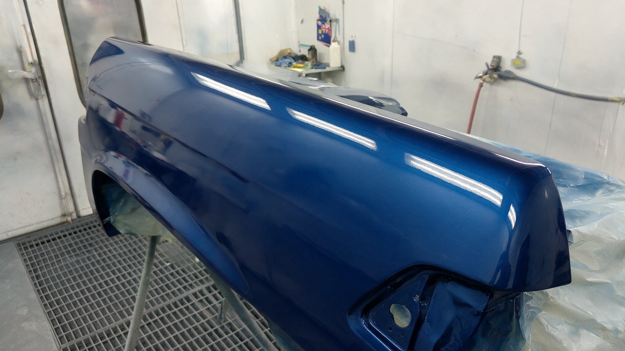 Car panel freshly sprayed in deep metallic blue inside a spray booth, glossy wet finish catching the even white booth lighting